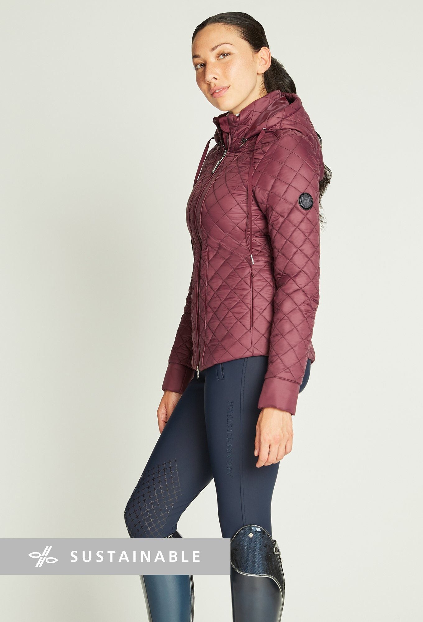 Woman wearing knee-length riding boots, blue riding leggings, maroon riding jacket.