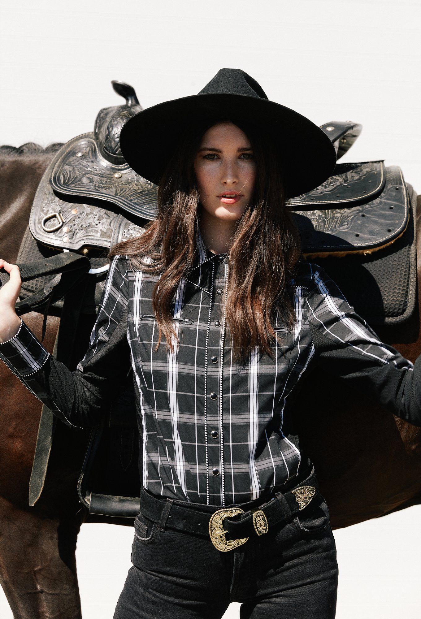 Woman wearing black and white check shirt and black riding pants with a black wide-brim hat.